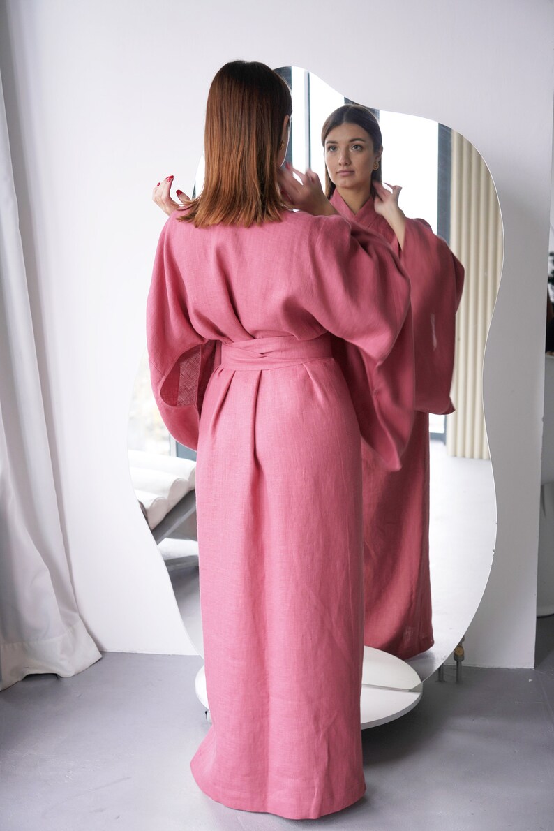 May include: A long, pink linen robe with a wide sash and long sleeves. The robe is tied at the waist and has a relaxed fit.