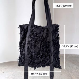 Peut inclure: Sac fourre-tout noir avec un motif en tissu d&eacute;chiquet&eacute; textur&eacute;. Le sac a de longues sangles et mesure 50 cm de large, 46 cm de haut, et les sangles mesurent 30 cm de long. Le sac est tenu par une main.