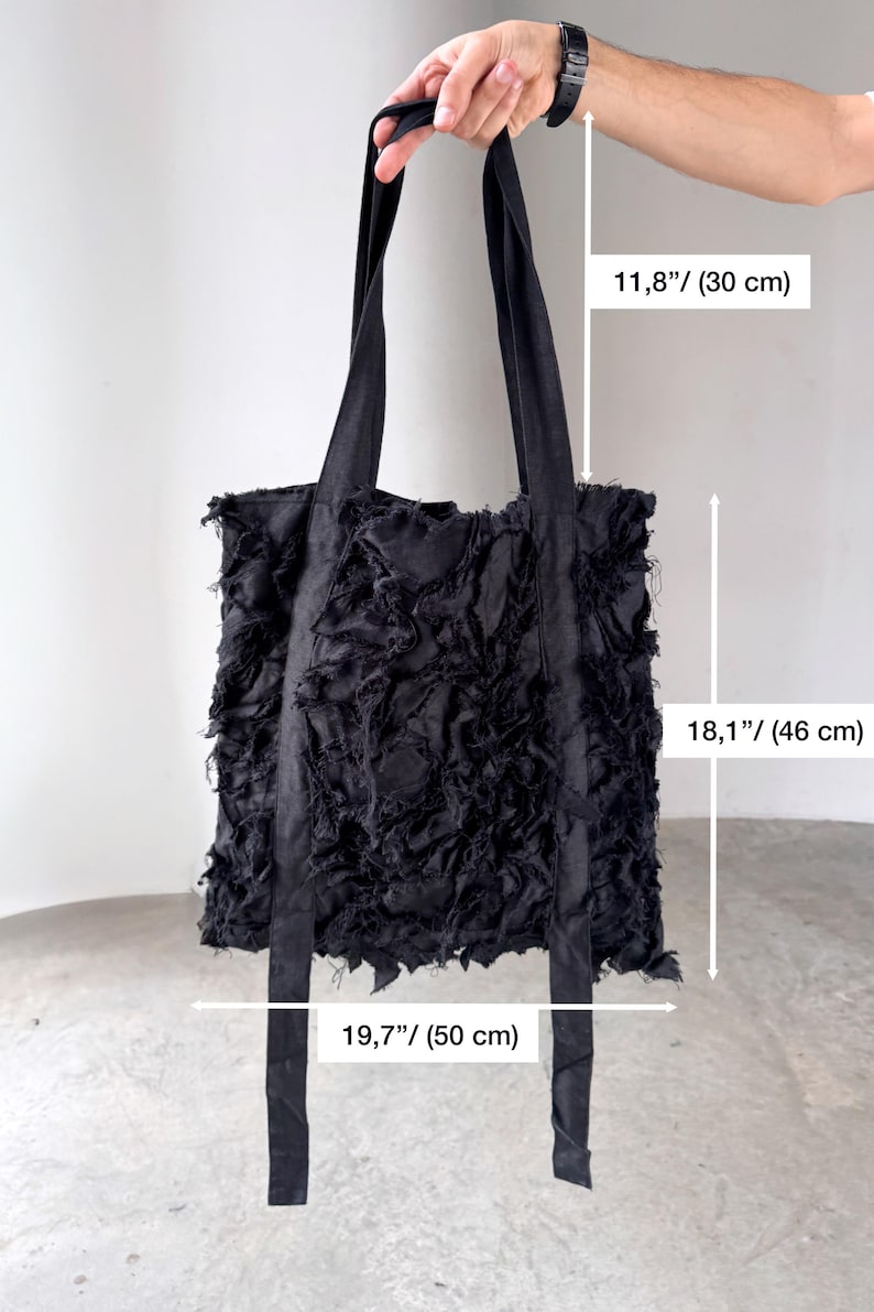 Peut inclure: Sac fourre-tout noir avec un motif en tissu d&eacute;chiquet&eacute; textur&eacute;. Le sac a de longues sangles et mesure 50 cm de large, 46 cm de haut, et les sangles mesurent 30 cm de long. Le sac est tenu par une main.