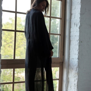 May include: A woman wearing a long, black linen robe stands in front of a window. The robe is sheer and has a flowing silhouette. The woman is looking down and her hair is long and dark.