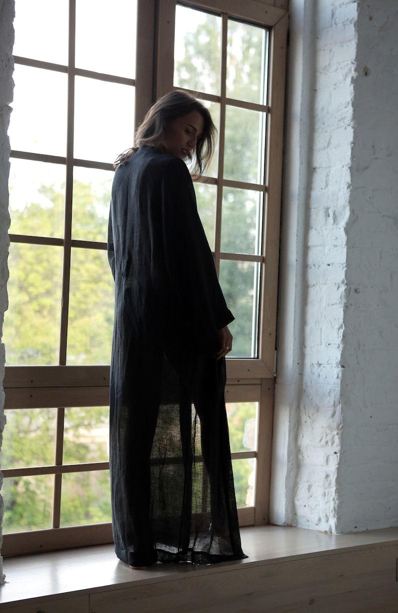 May include: A woman wearing a long, black linen robe stands in front of a window. The robe is sheer and has a flowing silhouette. The woman is looking down and her hair is long and dark.
