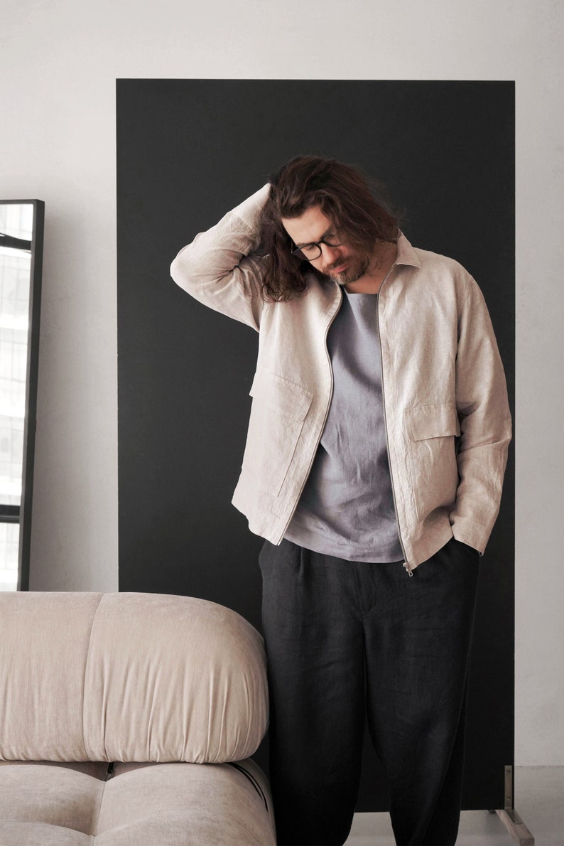 Puede incluir: Un hombre con una chaqueta de lino beige claro con cierre de cremallera, una camiseta azul claro y pantalones de lino negros. Est&aacute; de pie frente a un fondo negro.