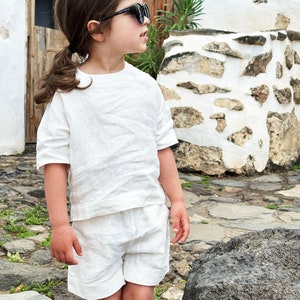 May include: A young person wearing a white linen outfit, including a short-sleeved shirt and shorts, with black sunglasses. The person is standing on a stone path in front of a white stone wall with the word "Jurdana" on a sign.