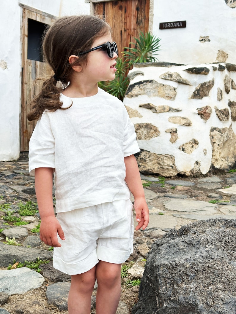 May include: A young person wearing a white linen outfit, including a short-sleeved shirt and shorts, with black sunglasses. The person is standing on a stone path in front of a white stone wall with the word "Jurdana" on a sign.