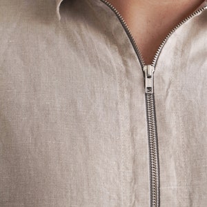 Puede incluir: Una camisa de lino beige con cierre de cremallera. La cremallera es plateada y tiene una peque&ntilde;a leng&uuml;eta de metal.