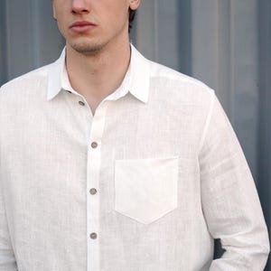 Pu&ograve; includere: Una camicia bianca di lino con colletto abbottonato e una singola tasca sul petto. La camicia ha una trama tessuta e &egrave; sbottonata nella parte superiore.