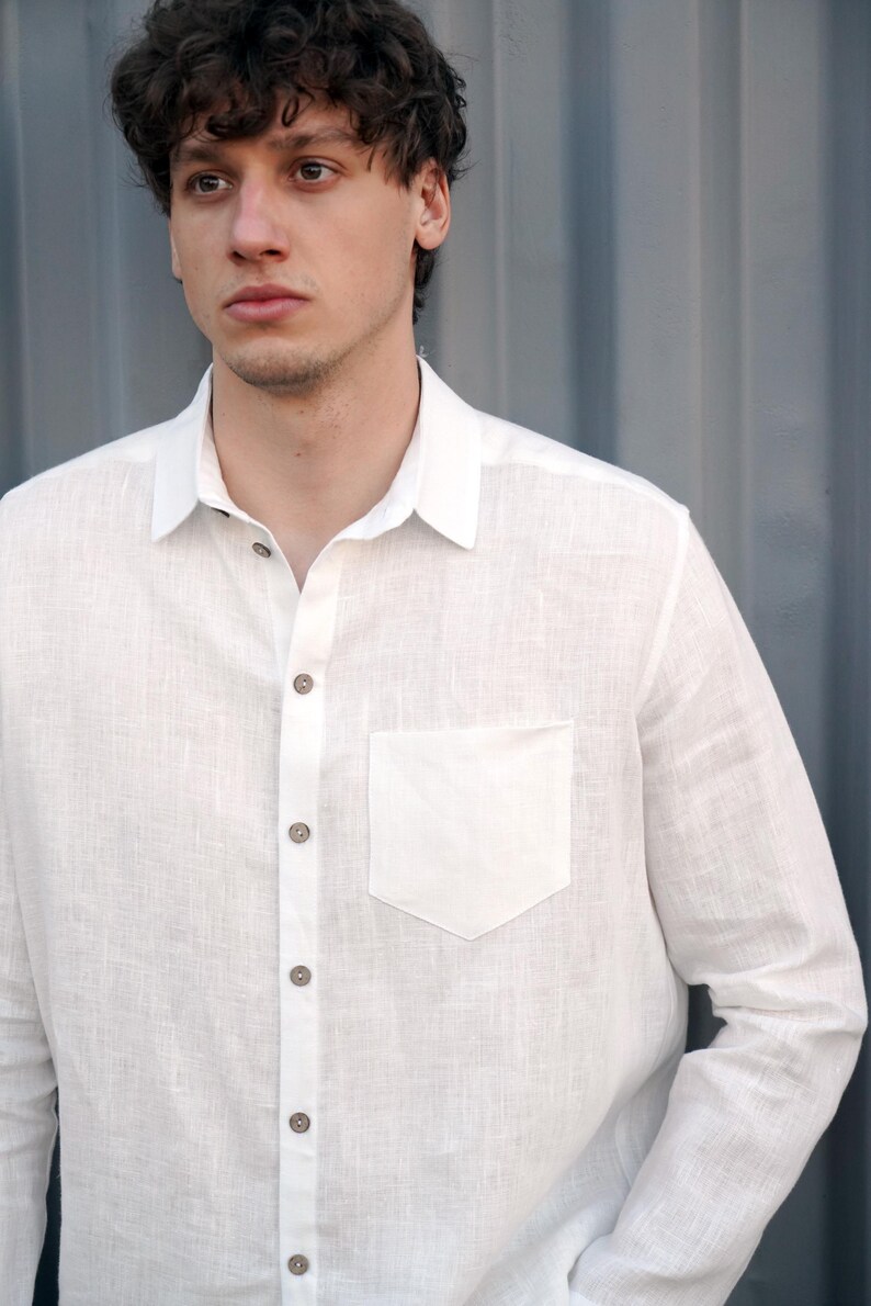 Pu&ograve; includere: Una camicia bianca di lino con colletto abbottonato e una singola tasca sul petto. La camicia ha una trama tessuta e &egrave; sbottonata nella parte superiore.