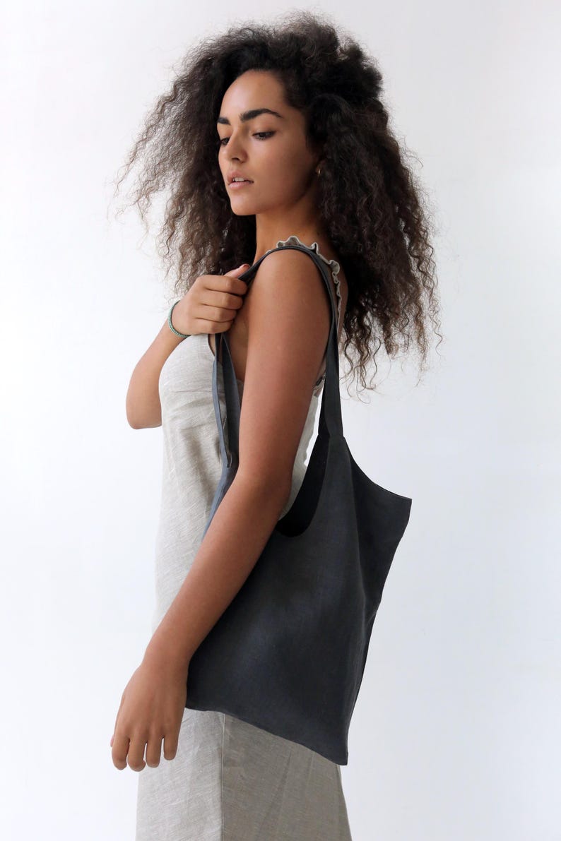 May include: A woman wearing a light grey dress holds a dark grey linen tote bag with a single strap.