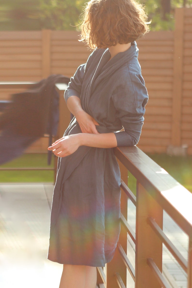 May include: A woman wearing a blue linen robe stands by a wooden railing. The robe has a relaxed fit and a shawl collar. The woman's hair is curly and brown.