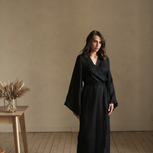 May include: A woman wearing a long black linen robe with a tie belt. The robe has wide sleeves and a V-neckline. She is standing on a wooden floor in front of a beige wall.