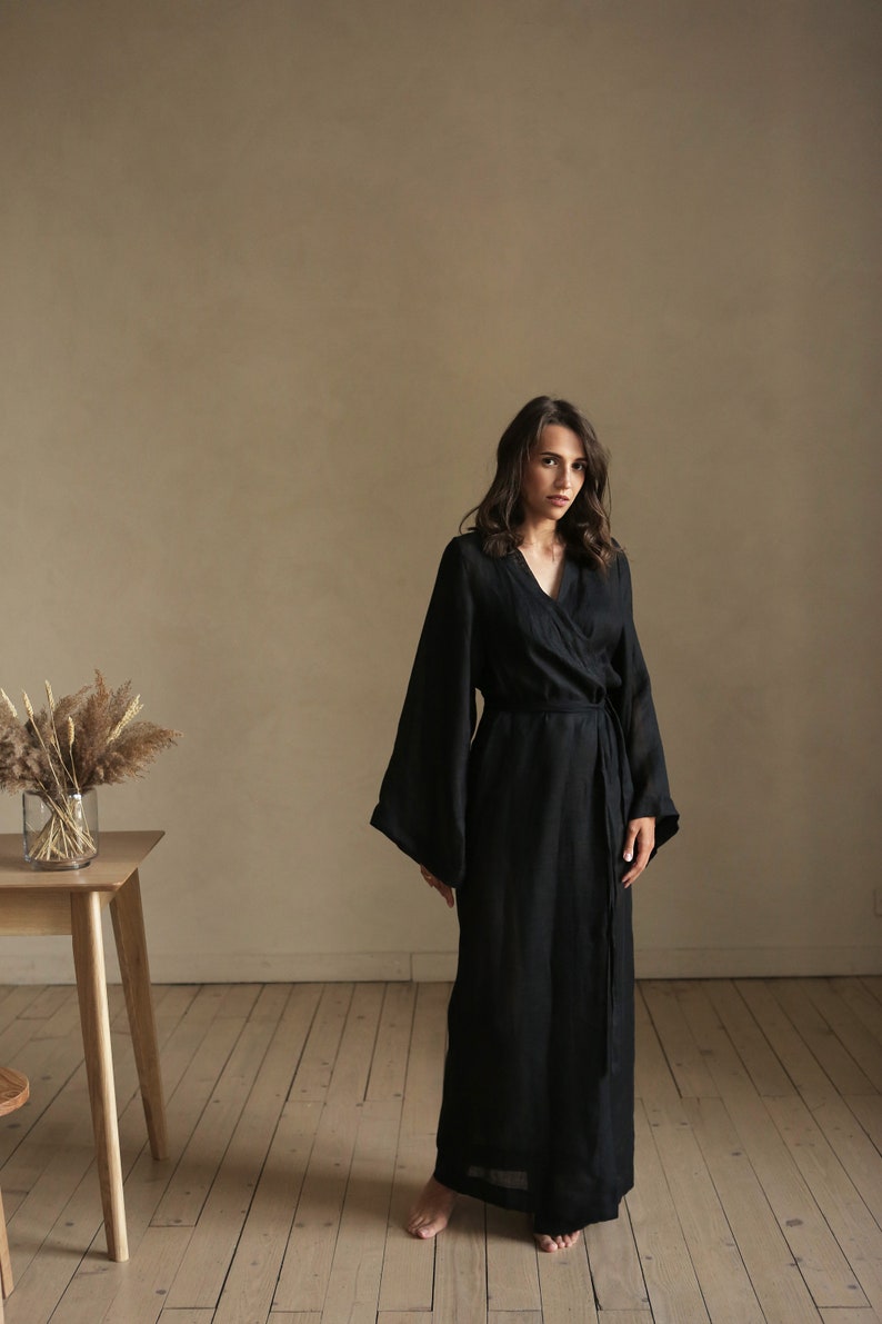 May include: A woman wearing a long black linen robe with a tie belt. The robe has wide sleeves and a V-neckline. She is standing on a wooden floor in front of a beige wall.