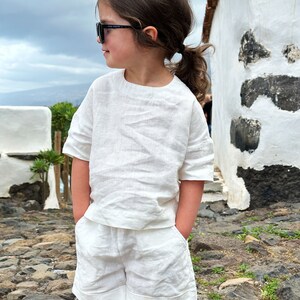 May include: A young person wearing a white linen outfit, including a short-sleeved top and shorts. They are wearing sunglasses and have their hair pulled back in a ponytail.