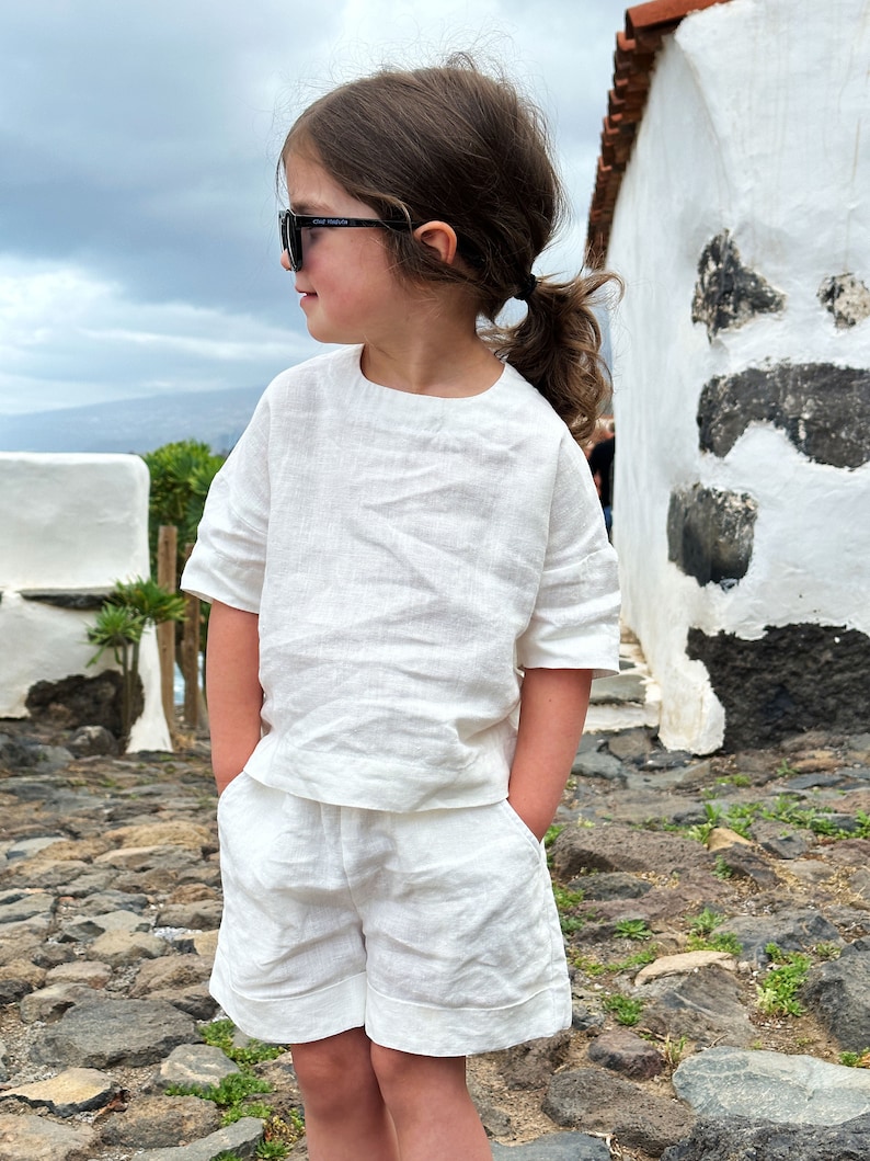 May include: A young person wearing a white linen outfit, including a short-sleeved top and shorts. They are wearing sunglasses and have their hair pulled back in a ponytail.