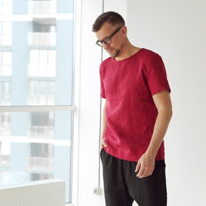 May include: A man wearing a red linen t-shirt and black pants. He is standing in front of a window.