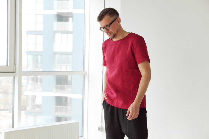 May include: A man wearing a red linen t-shirt and black pants. He is standing in front of a window.