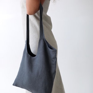 May include: A dark blue linen tote bag with a long strap. The bag is being held by a person wearing a light grey dress.