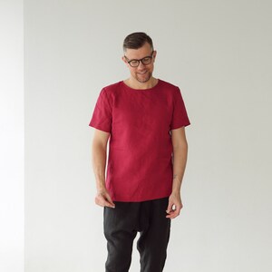 May include: A man wearing a red linen shirt with short sleeves and black pants. He is smiling and looking down.