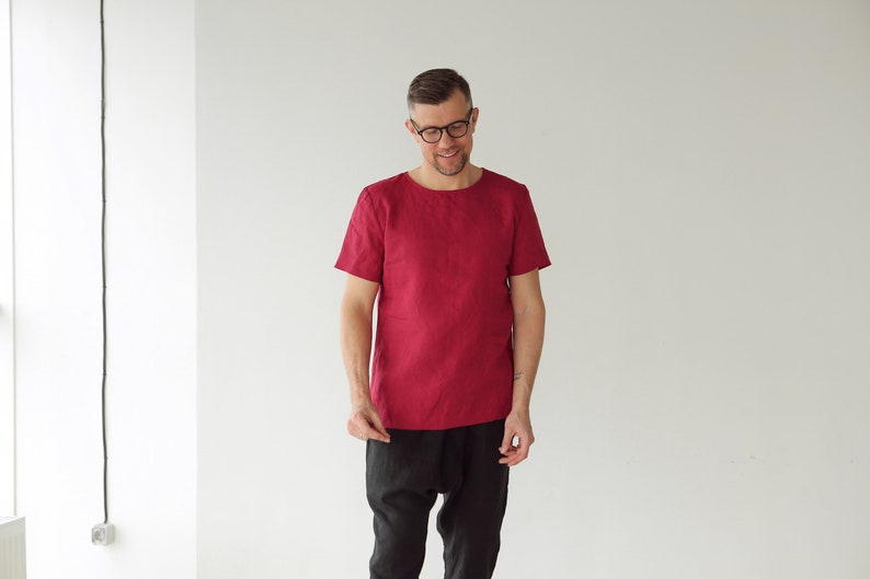 May include: A man wearing a red linen shirt with short sleeves and black pants. He is smiling and looking down.