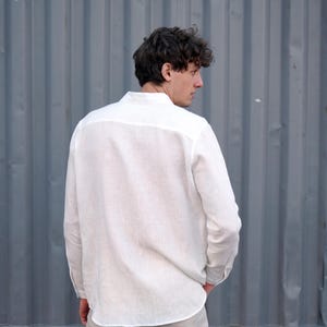 Pu&ograve; includere: Una camicia bianca di lino a maniche lunghe e colletto abbottonato. La camicia &egrave; indossata da una persona in piedi davanti a un muro di metallo ondulato grigio.