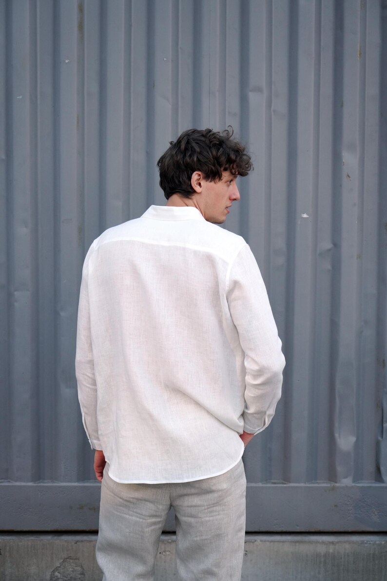 Pu&ograve; includere: Una camicia bianca di lino a maniche lunghe e colletto abbottonato. La camicia &egrave; indossata da una persona in piedi davanti a un muro di metallo ondulato grigio.