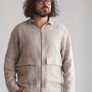 Puede incluir: Una chaqueta de lino beige claro con cierre de cremallera y un bolsillo grande en la parte delantera. La chaqueta tiene mangas largas y cuello.