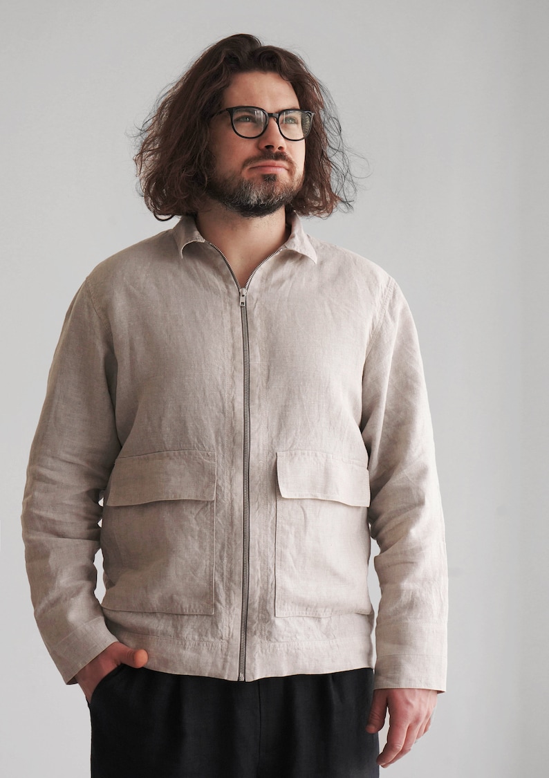 Puede incluir: Una chaqueta de lino beige claro con cierre de cremallera y un bolsillo grande en la parte delantera. La chaqueta tiene mangas largas y cuello.