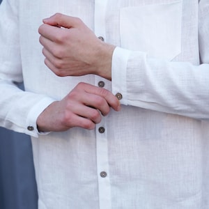 Pu&ograve; includere: Una camicia bianca di lino con colletto abbottonato e una sola tasca sul petto. La camicia &egrave; sbottonata al polsino e i bottoni sono marroni.