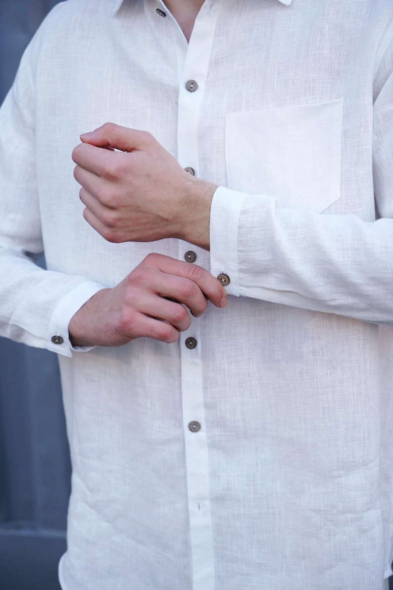 Pu&ograve; includere: Una camicia bianca di lino con colletto abbottonato e una sola tasca sul petto. La camicia &egrave; sbottonata al polsino e i bottoni sono marroni.