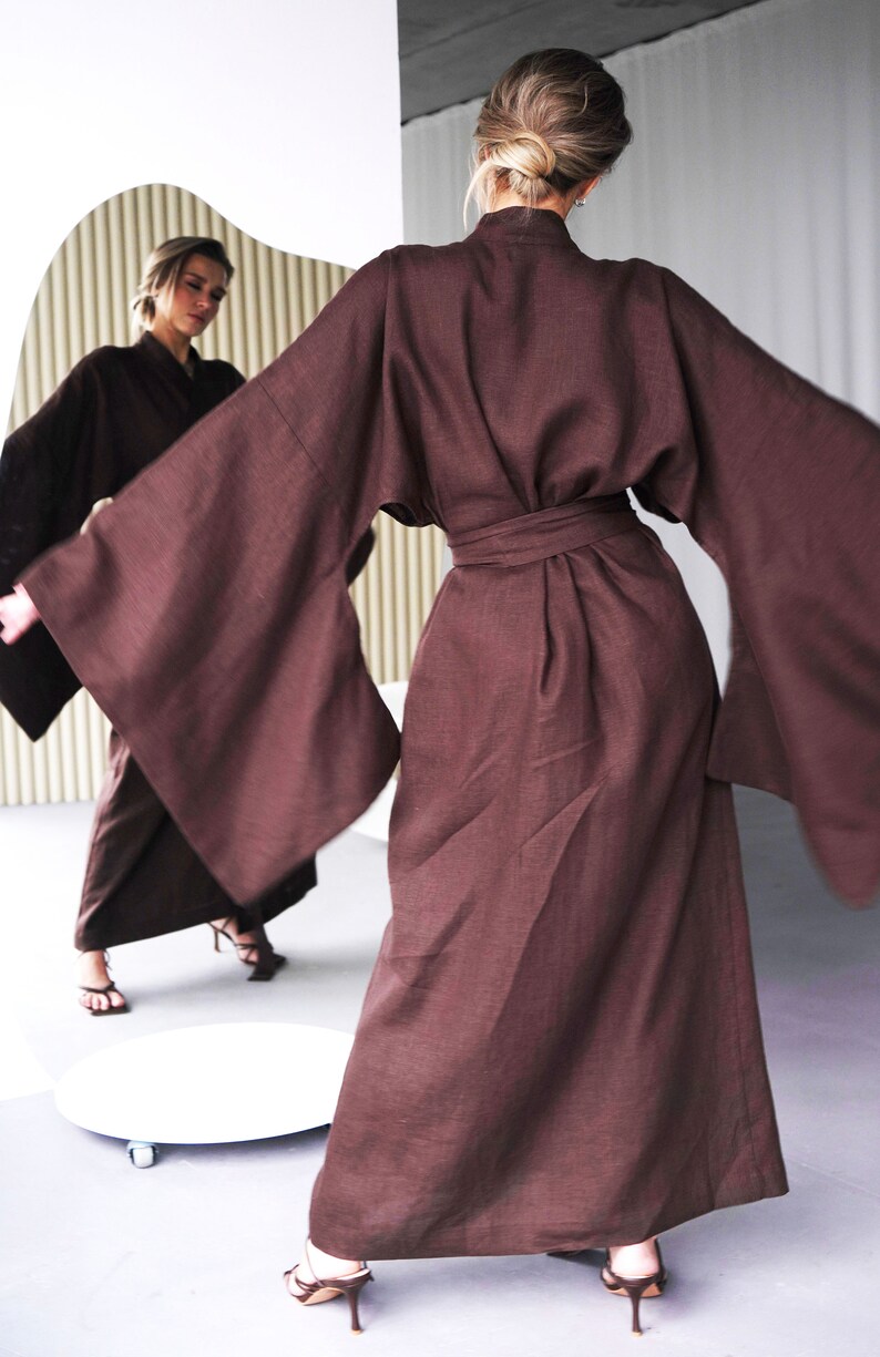 May include: A long, brown linen robe with wide sleeves and a sash tie at the waist. The robe is worn by a person standing in front of a mirror.