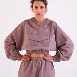 May include: A mauve linen hoodie with a drawstring hood and a large pocket on the front. The hoodie is cropped and has elastic at the waist. The model is also wearing matching mauve linen pants with cargo pockets.