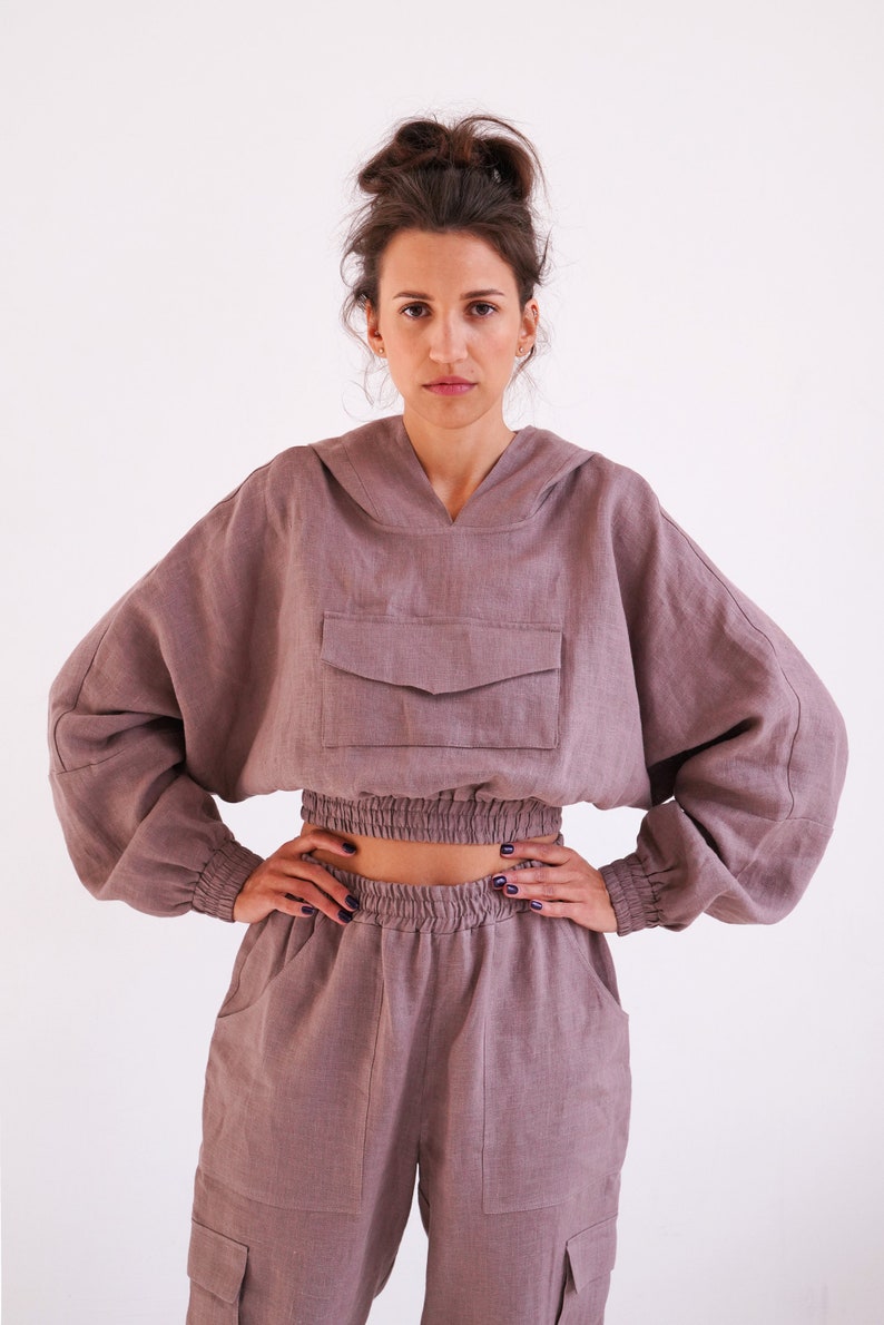 May include: A mauve linen hoodie with a drawstring hood and a large pocket on the front. The hoodie is cropped and has elastic at the waist. The model is also wearing matching mauve linen pants with cargo pockets.