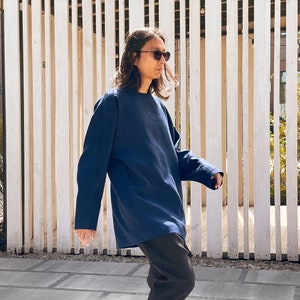 May include: A person wearing a dark blue long-sleeved shirt and black pants, standing in front of a wooden slat wall. They are wearing sunglasses and looking to the right.