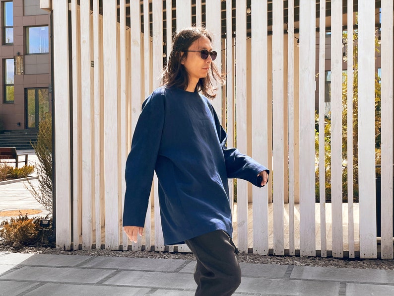 May include: A person wearing a dark blue long-sleeved shirt and black pants, standing in front of a wooden slat wall. They are wearing sunglasses and looking to the right.