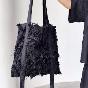 Peut inclure: Sac fourre-tout noir avec de longues sangles. Le corps du sac pr&eacute;sente un design textur&eacute; et effiloch&eacute;. Les sangles sont lisses et solides. Le sac est tenu par une main, et l'arri&egrave;re-plan est un mur blanc uni.