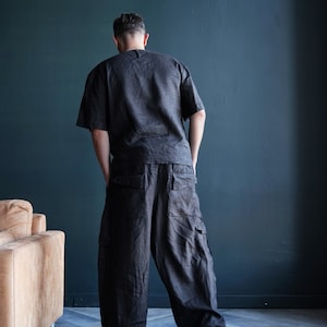 Pu&ograve; includere: Un completo in lino grigio scuro, composto da un top a maniche corte e pantaloni cargo a gamba larga. I pantaloni hanno ampie tasche laterali. L'outfit &egrave; indossato con scarpe da ginnastica scure. Lo sfondo &egrave; un muro blu scuro.