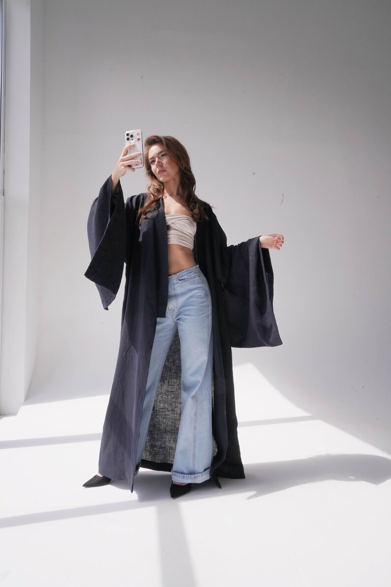 May include: A woman wearing a long, dark blue robe with wide sleeves and light blue jeans. She is holding a smartphone in her right hand and looking at the camera.