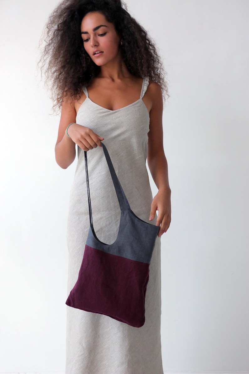 May include: A woman wearing a light grey linen dress holds a two-toned tote bag. The bag is made of a dark grey fabric on the top half and a burgundy fabric on the bottom half.