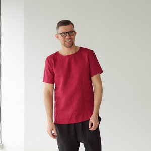 May include: A man wearing a red linen shirt with short sleeves and black pants. He is smiling and looking at the camera.