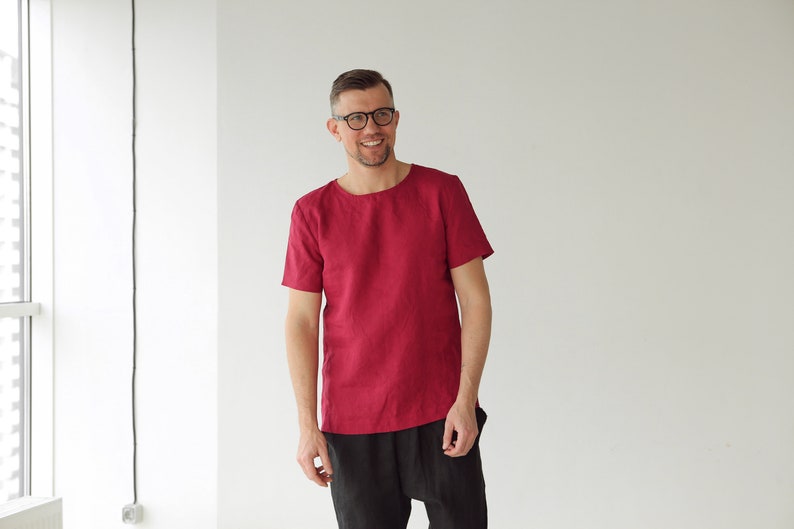May include: A man wearing a red linen shirt with short sleeves and black pants. He is smiling and looking at the camera.