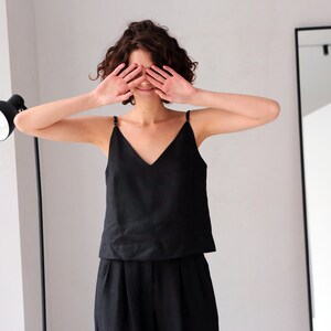 以下が含まれることがあります： 黒いノースリーブトップと黒いパンツを着用した女性。彼女は両手で目を覆い、笑顔を見せています。