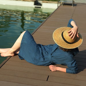 May include: A woman wearing a blue linen dress and a wide-brimmed straw hat relaxes on a wooden dock by a pool.