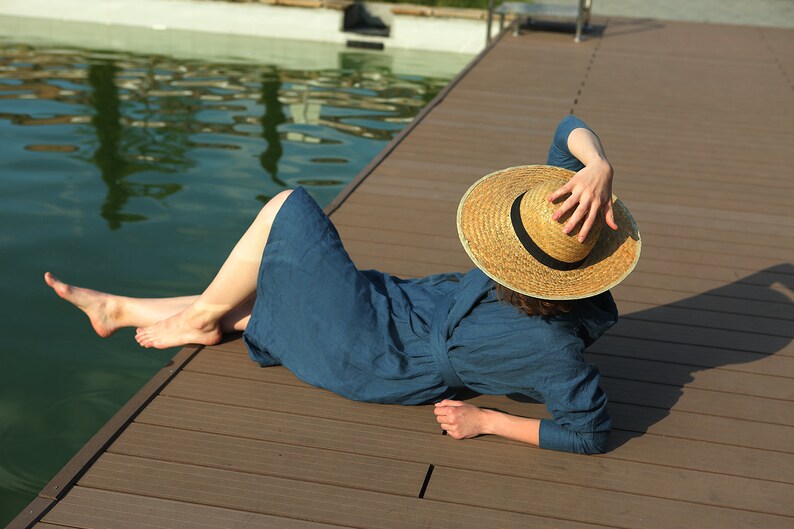May include: A woman wearing a blue linen dress and a wide-brimmed straw hat relaxes on a wooden dock by a pool.