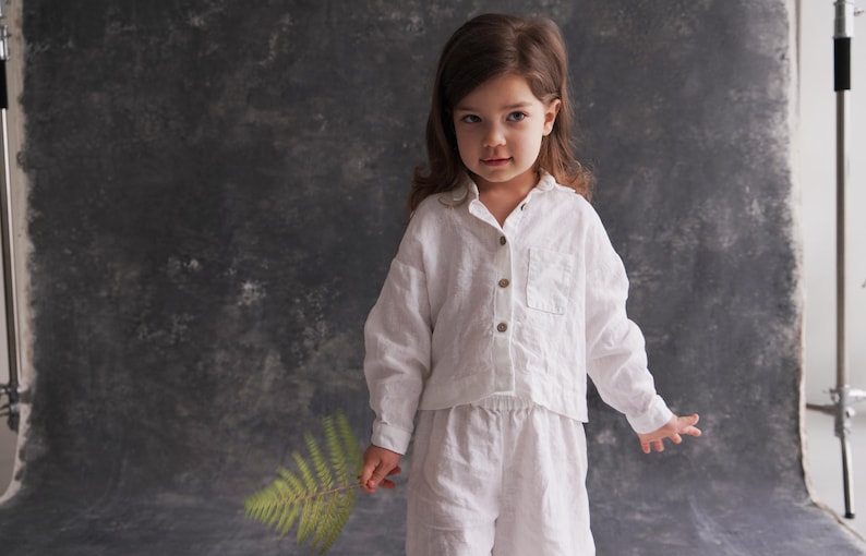 Pu&ograve; includere: Una bambina con una camicia e pantaloni di lino bianco sta in piedi davanti a uno sfondo grigio testurizzato. Tiene un'edera verde nella mano.