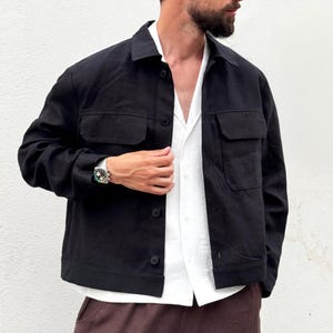 May include: A black button-down jacket featuring a classic collar, long sleeves, and two chest pockets with flaps. The jacket is worn over a white collared shirt, paired with dark brown trousers, and accessorised with a silver-toned wristwatch.