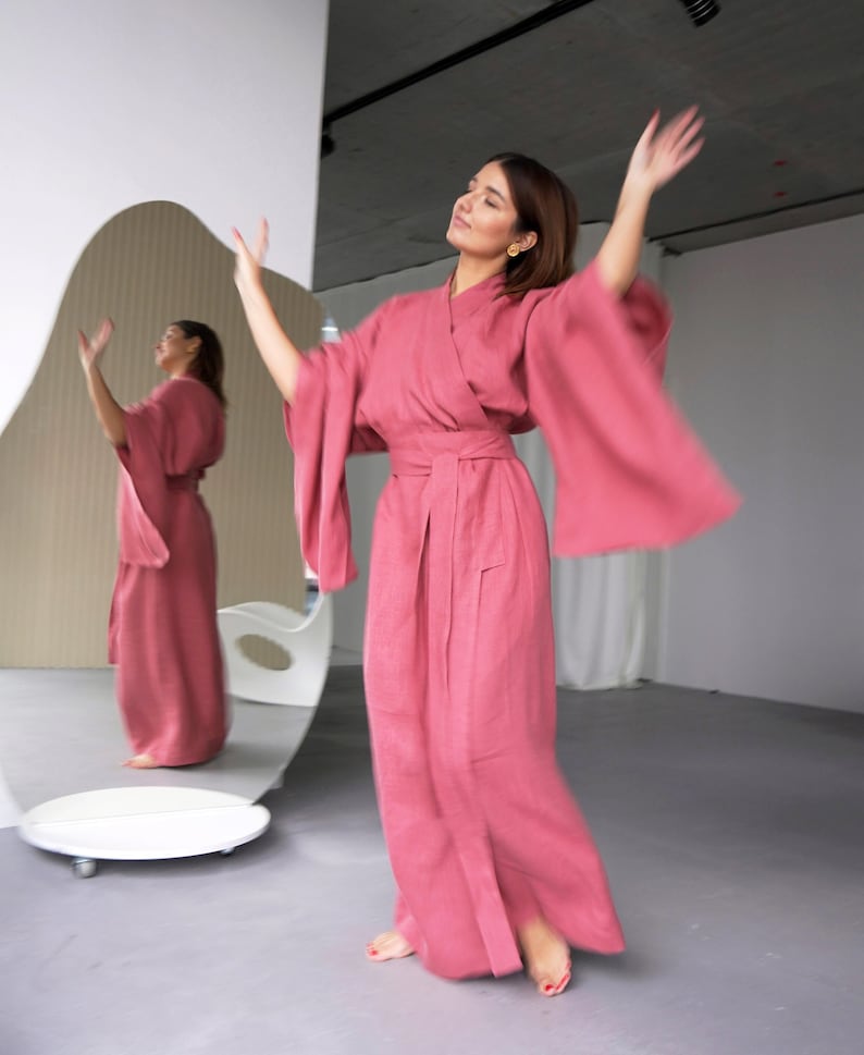 May include: A woman wearing a long, pink linen robe with wide sleeves and a sash tied at the waist. She is standing in a room with a large, white mirror behind her.
