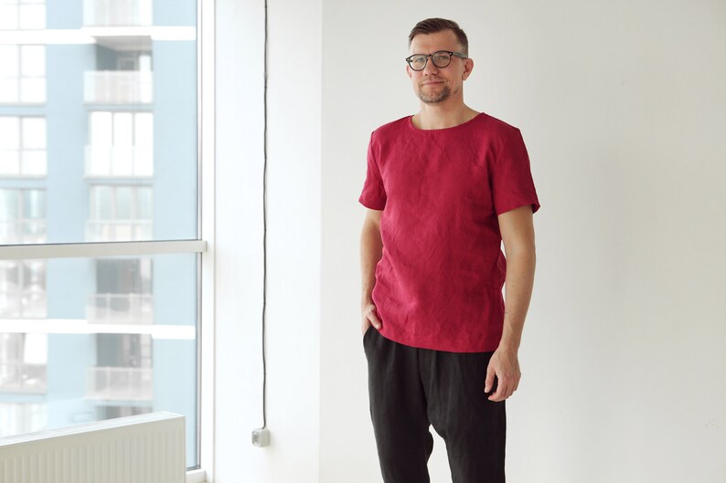 May include: A man wearing a red linen shirt with a round neckline and black linen pants. He is standing in front of a window.