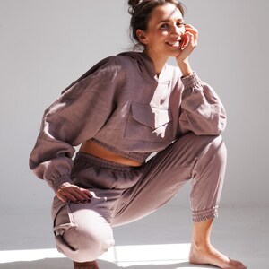 May include: A woman wearing a mauve-colored linen hoodie and matching pants. The hoodie has a large pocket on the front and the pants have a drawstring waist.