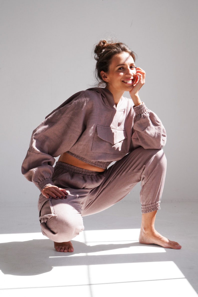 May include: A woman wearing a mauve-colored linen hoodie and matching pants. The hoodie has a large pocket on the front and the pants have a drawstring waist.