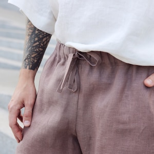 Pu&ograve; includere: Primo piano di una persona che indossa una camicia bianca a maniche corte e pantaloni di lino rosa polveroso. I pantaloni hanno una vita con coulisse. La persona ha un tatuaggio sul braccio.