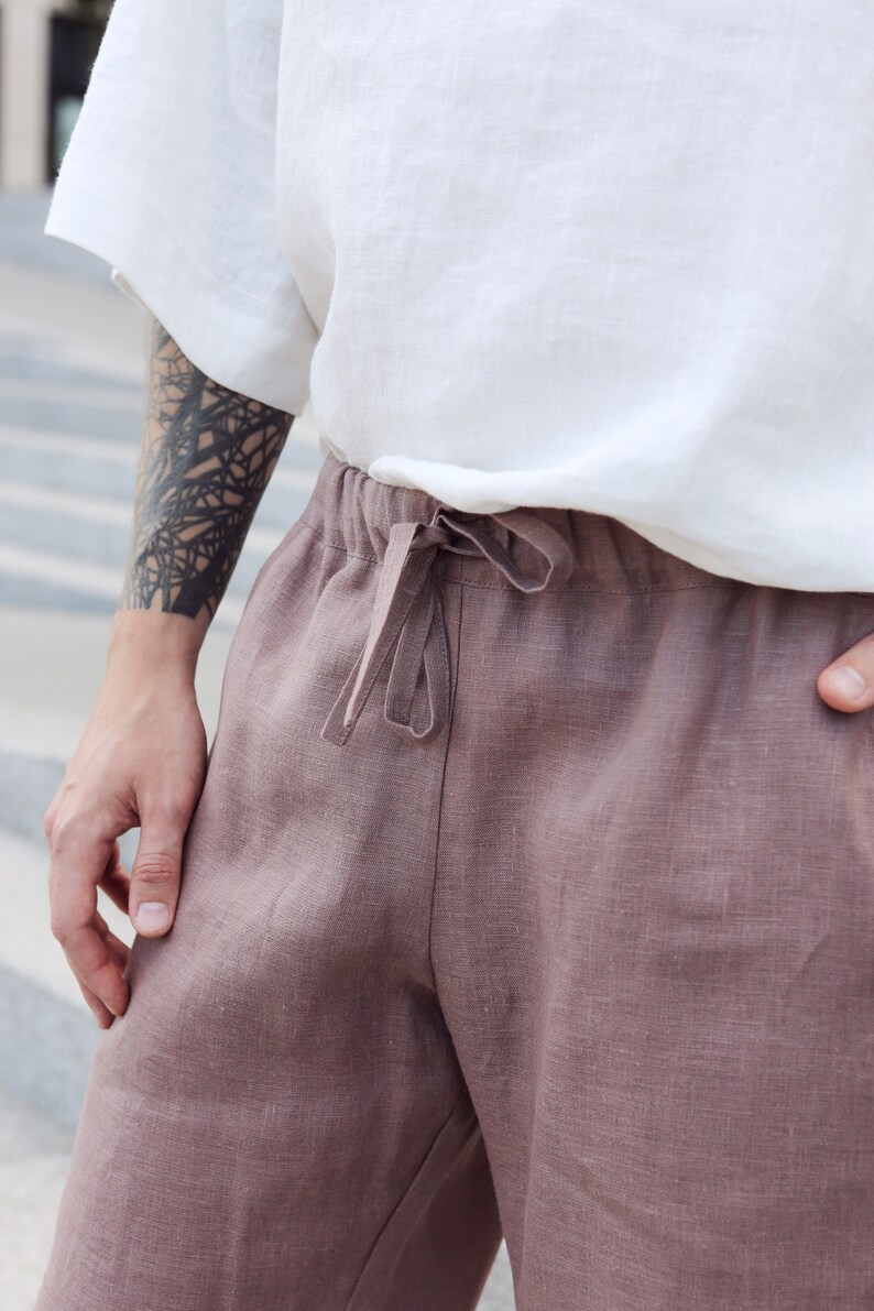 Pu&ograve; includere: Primo piano di una persona che indossa una camicia bianca a maniche corte e pantaloni di lino rosa polveroso. I pantaloni hanno una vita con coulisse. La persona ha un tatuaggio sul braccio.